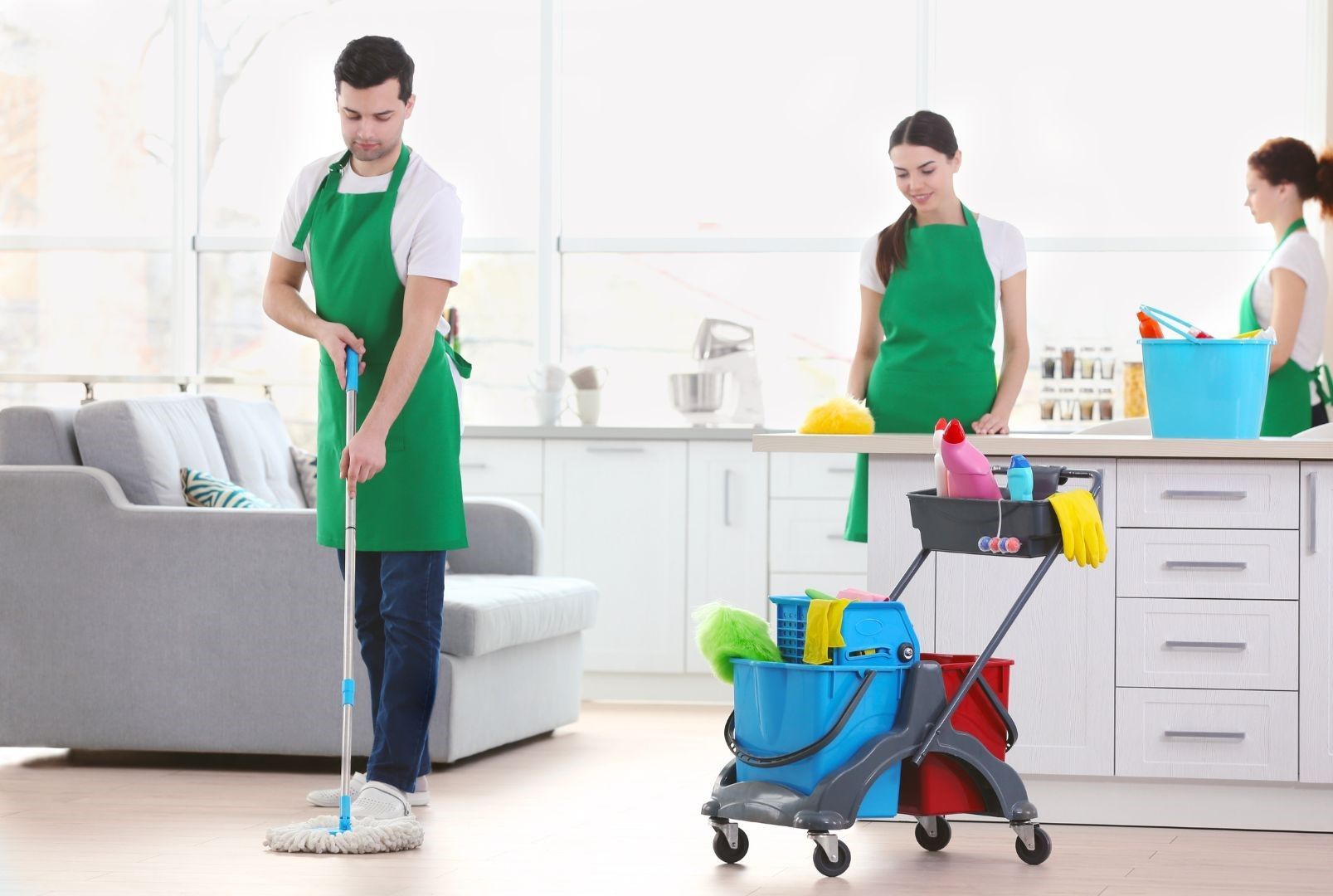 A man is mopping the floor in a living room while two cleaners are cleaning the kitchen.