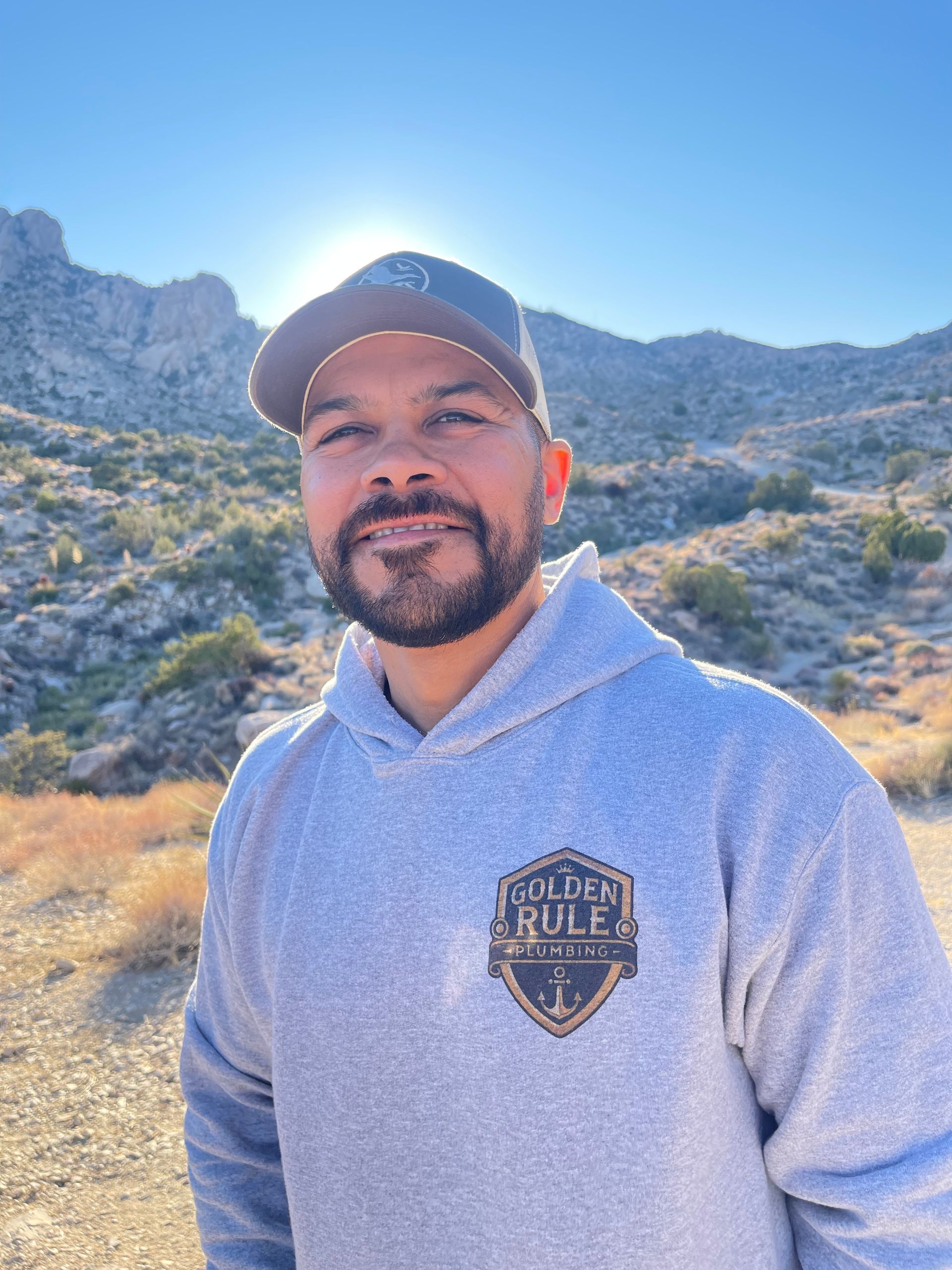 A man with a beard wearing a hat and a hoodie is standing in front of a mountain.
