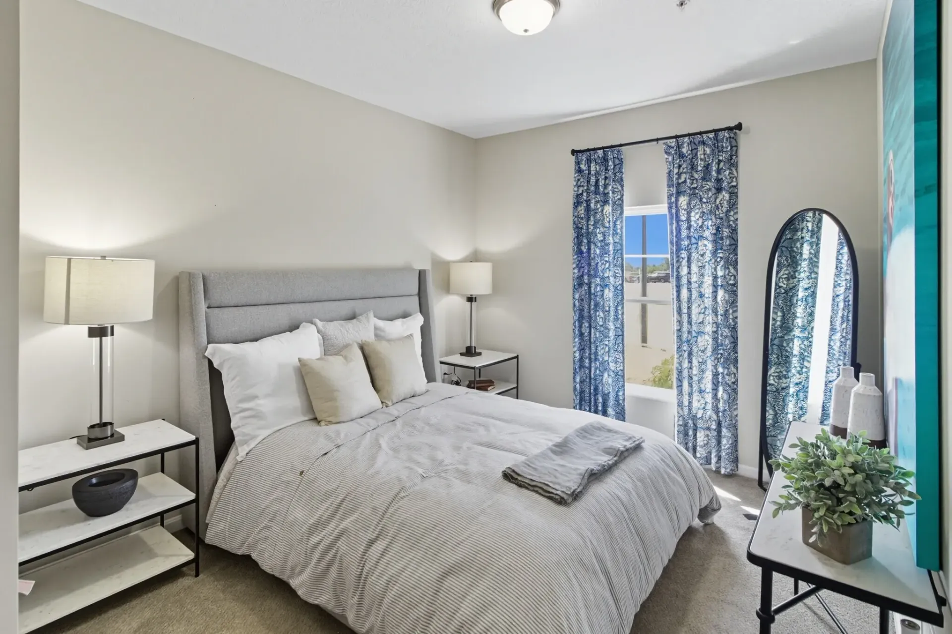 A neutrally decorated bedroom featuring a grey upholstered headboard, patterned bedding, blue curtains, and side tables.