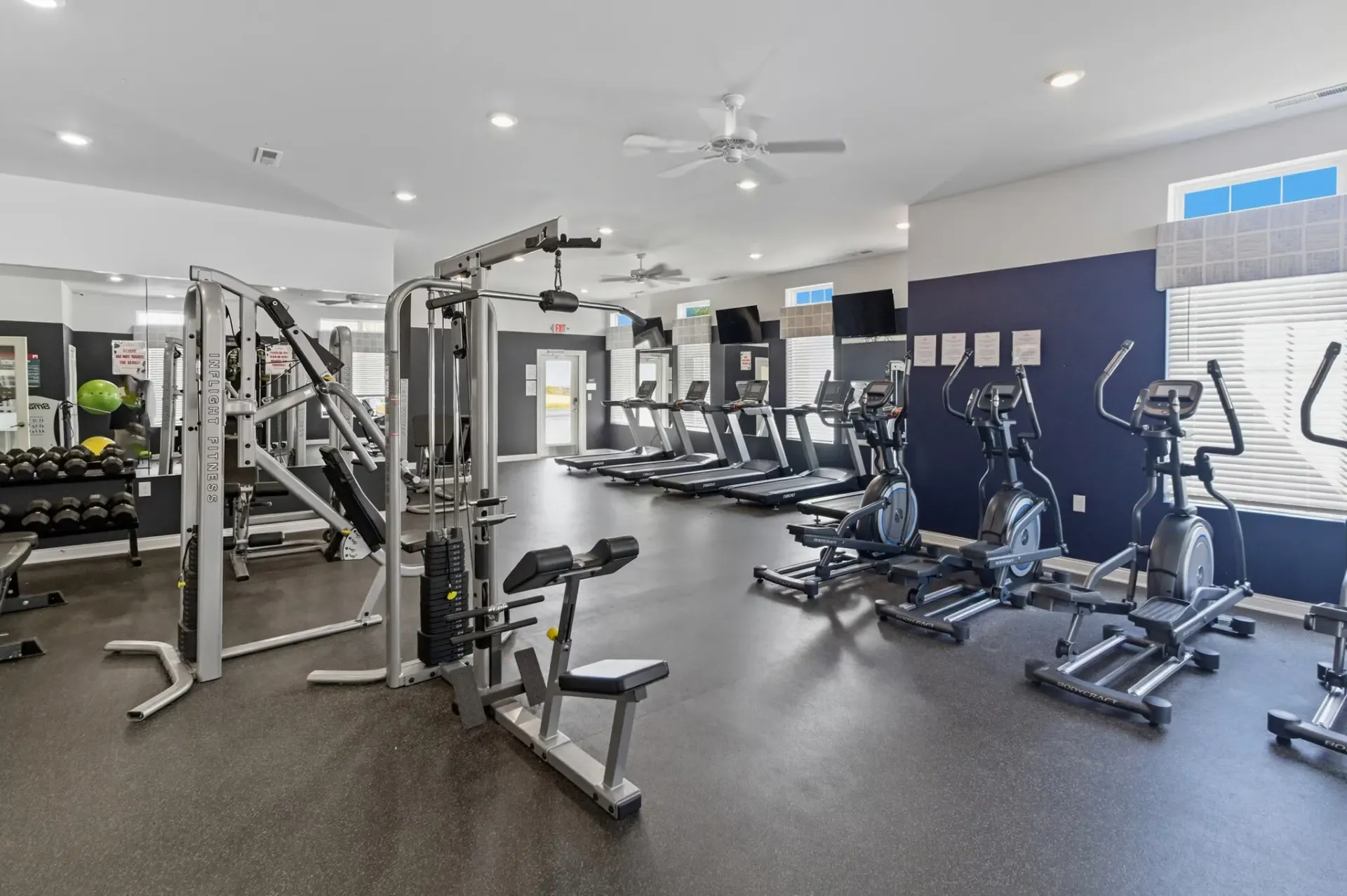 A brightly lit fitness center featuring rows of elliptical machines, a multi-station weight gym, and a rack of dumbbells.