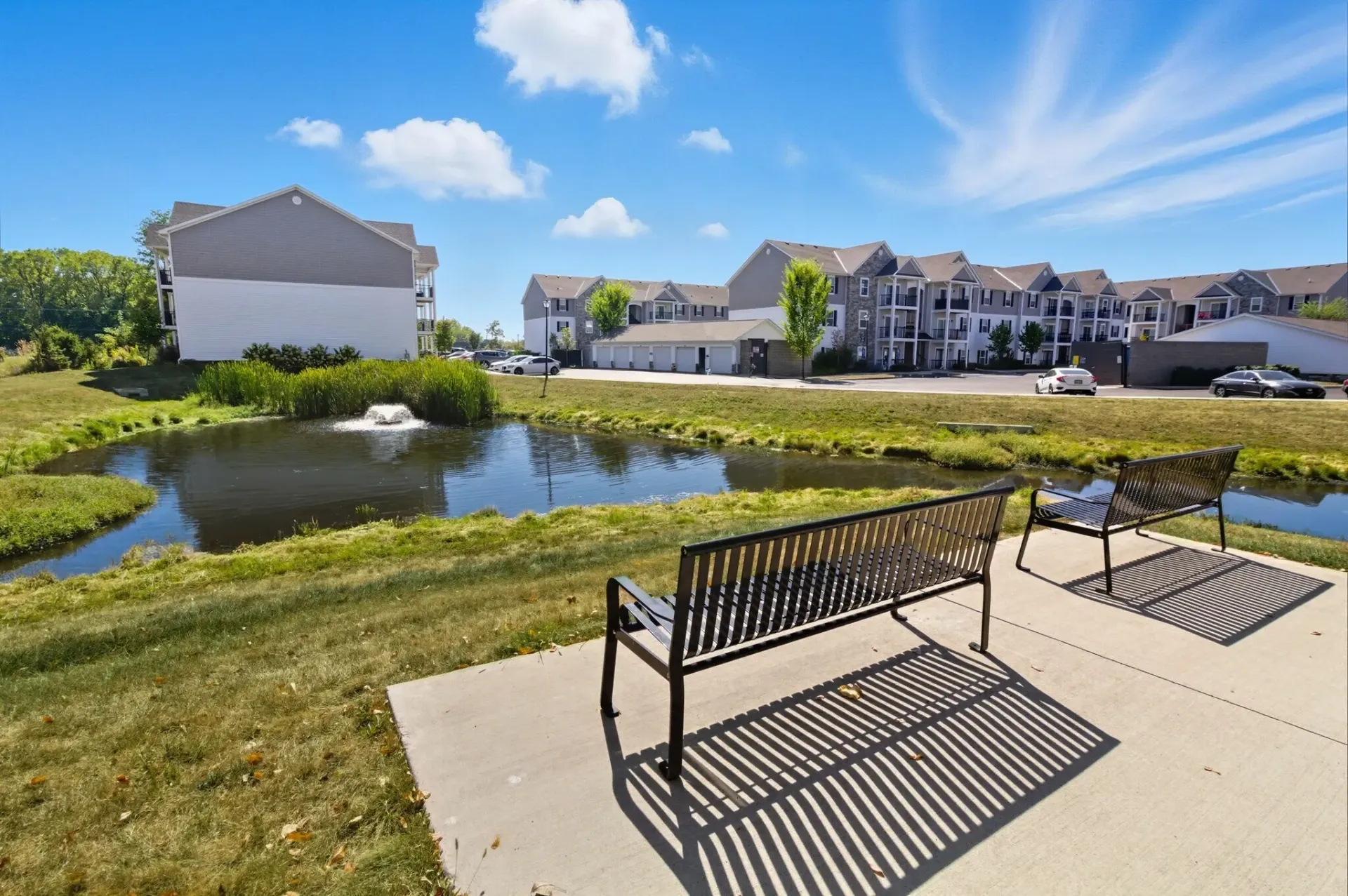 Two metal park benches overlook a small pond with a fountain in front of multi-story apartment buildings under a blue sky.