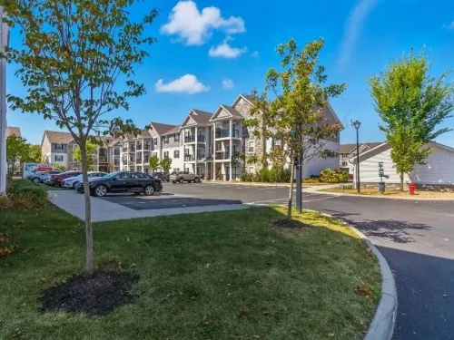 A multi-story residential apartment complex with a paved parking lot, green grass, and trees under a clear blue sky.