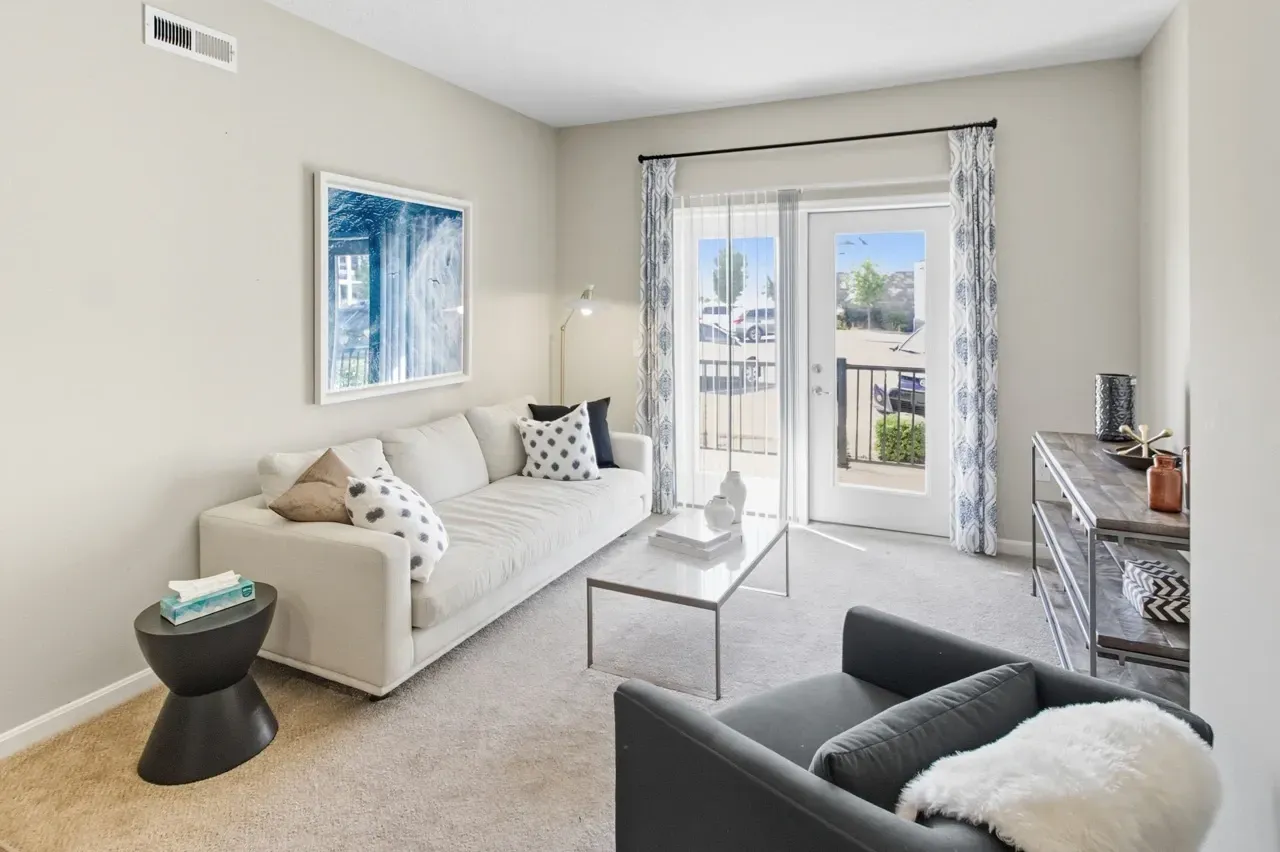 Modern living room with a white sofa, coffee table, and armchair, leading to a balcony with an outdoor view.