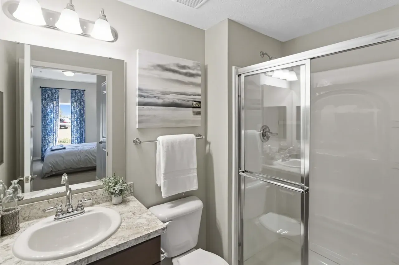 Bathroom with vanity, toilet, and glass-enclosed shower. Bedroom visible in mirror reflection.