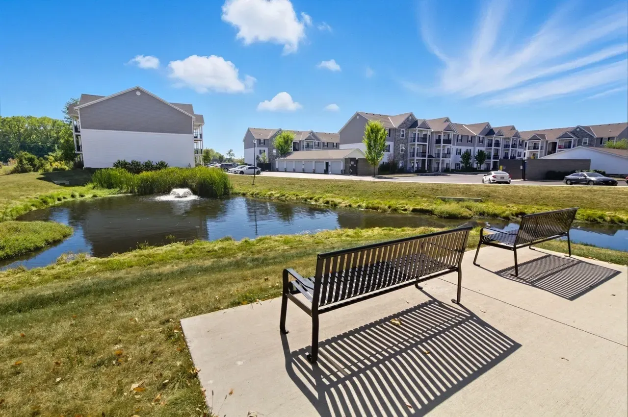 Outdoor area with a pond, fountain, benches, and apartment buildings.