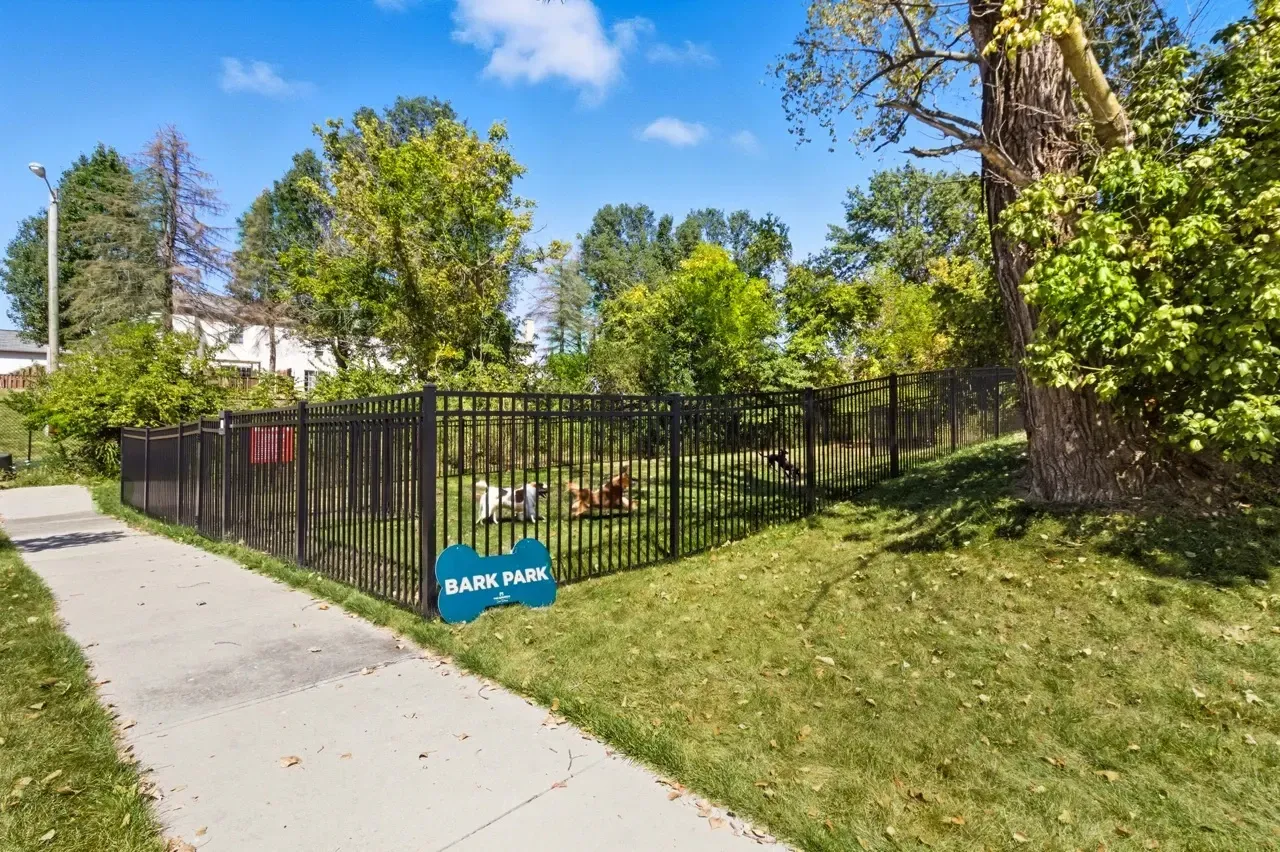 Fenced dog park with grass, trees, and a 'Bark Park' sign. Several dogs are playing inside.