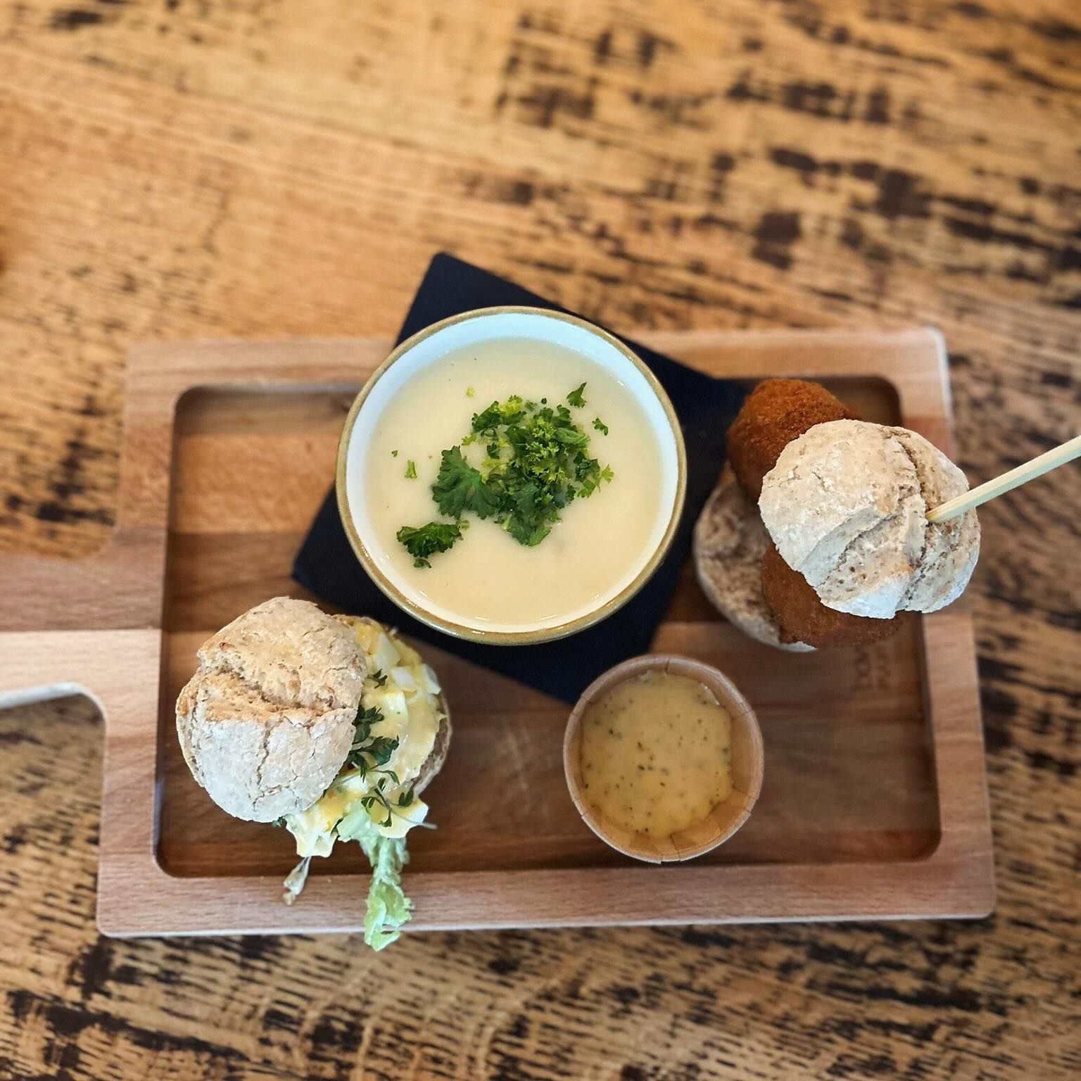 Een houten serveerplank met daarop een kommetje soep met kruiden, een mini-sandwich, gefrituurde snacks aan een spiesje en saus.