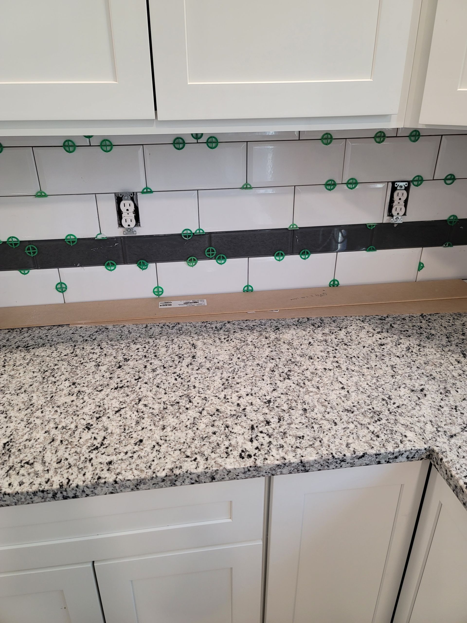 Backsplash tile installation in progress above a granite countertop, with green tile spacers visible.