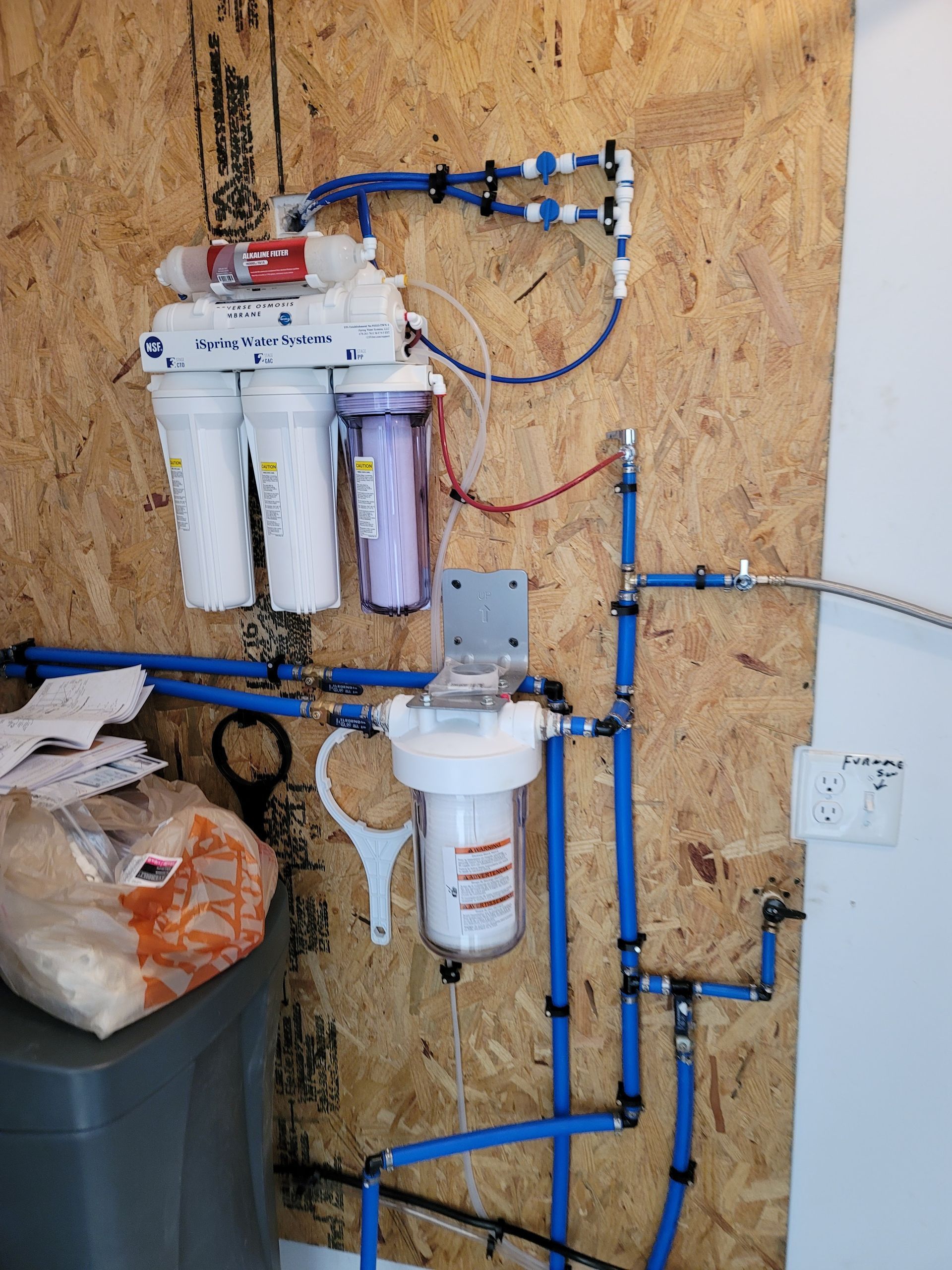 Water filtration system with blue pipes, white filters, and attached to wood paneling.