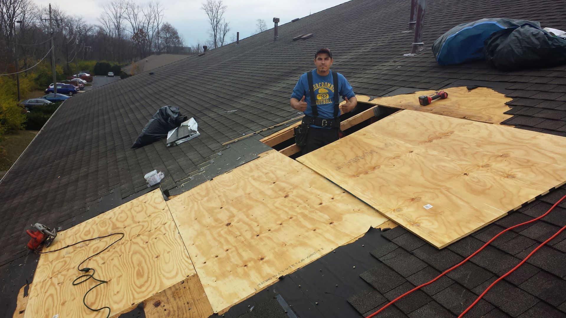 Man on a roof repairing wood panels. Black asphalt shingles. Outdoors, overcast.