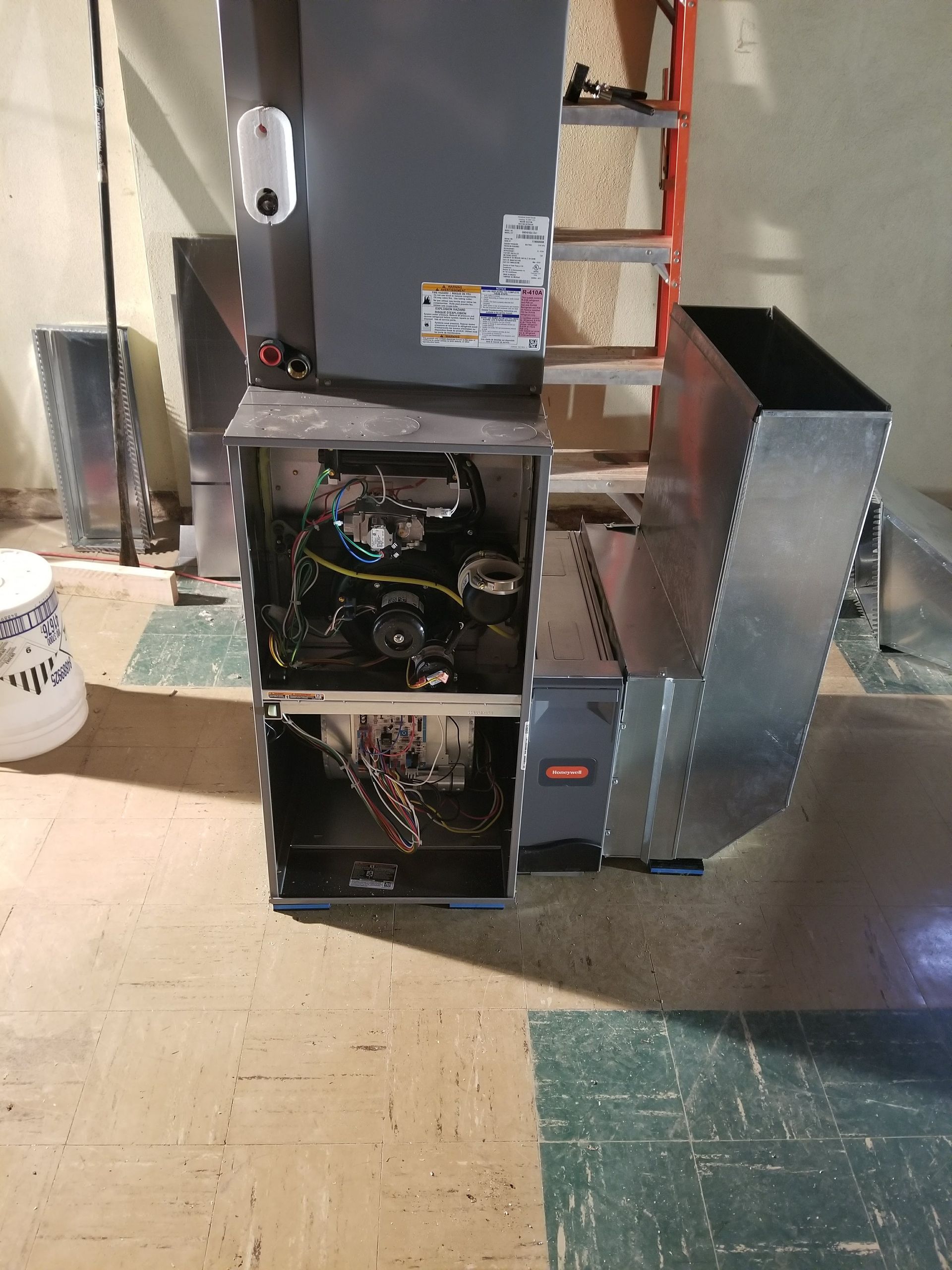 Furnace with exposed internal components, ducts, and a white bucket in a basement setting.