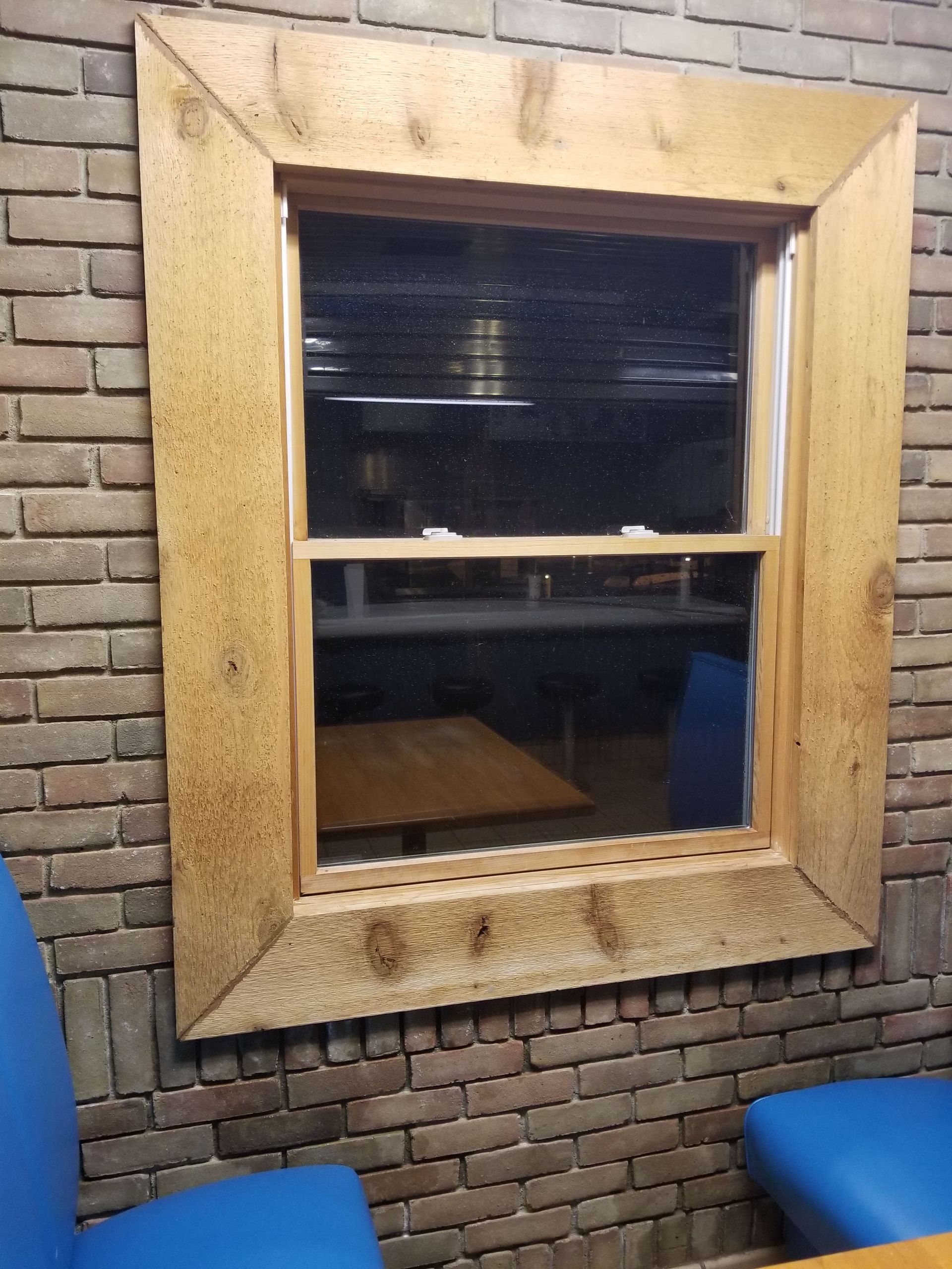 Wooden framed window on brick wall, reflecting a room with table and blue chairs.