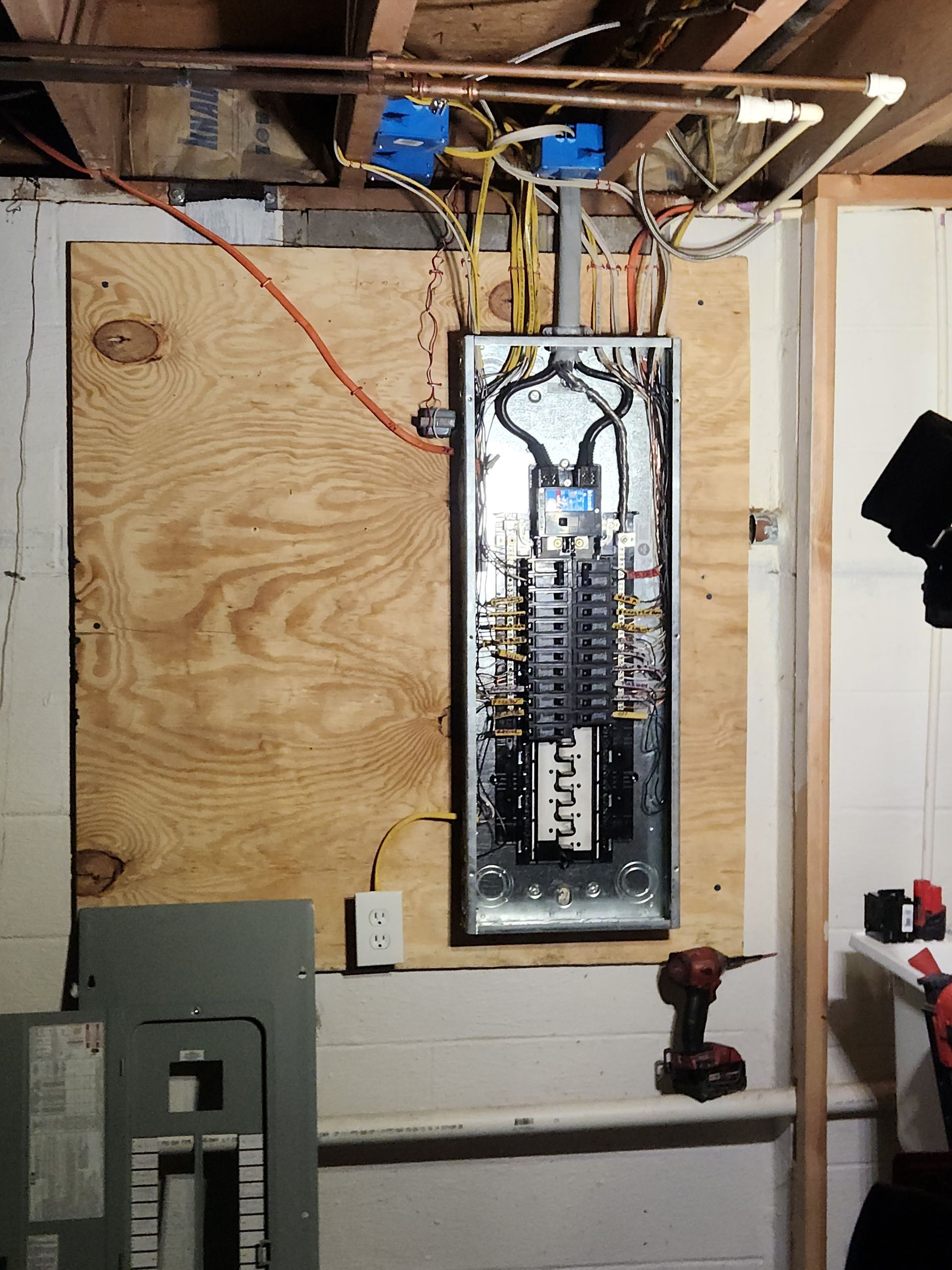Electrical panel mounted on plywood in a basement, wiring visible. A power drill and other tools are nearby.