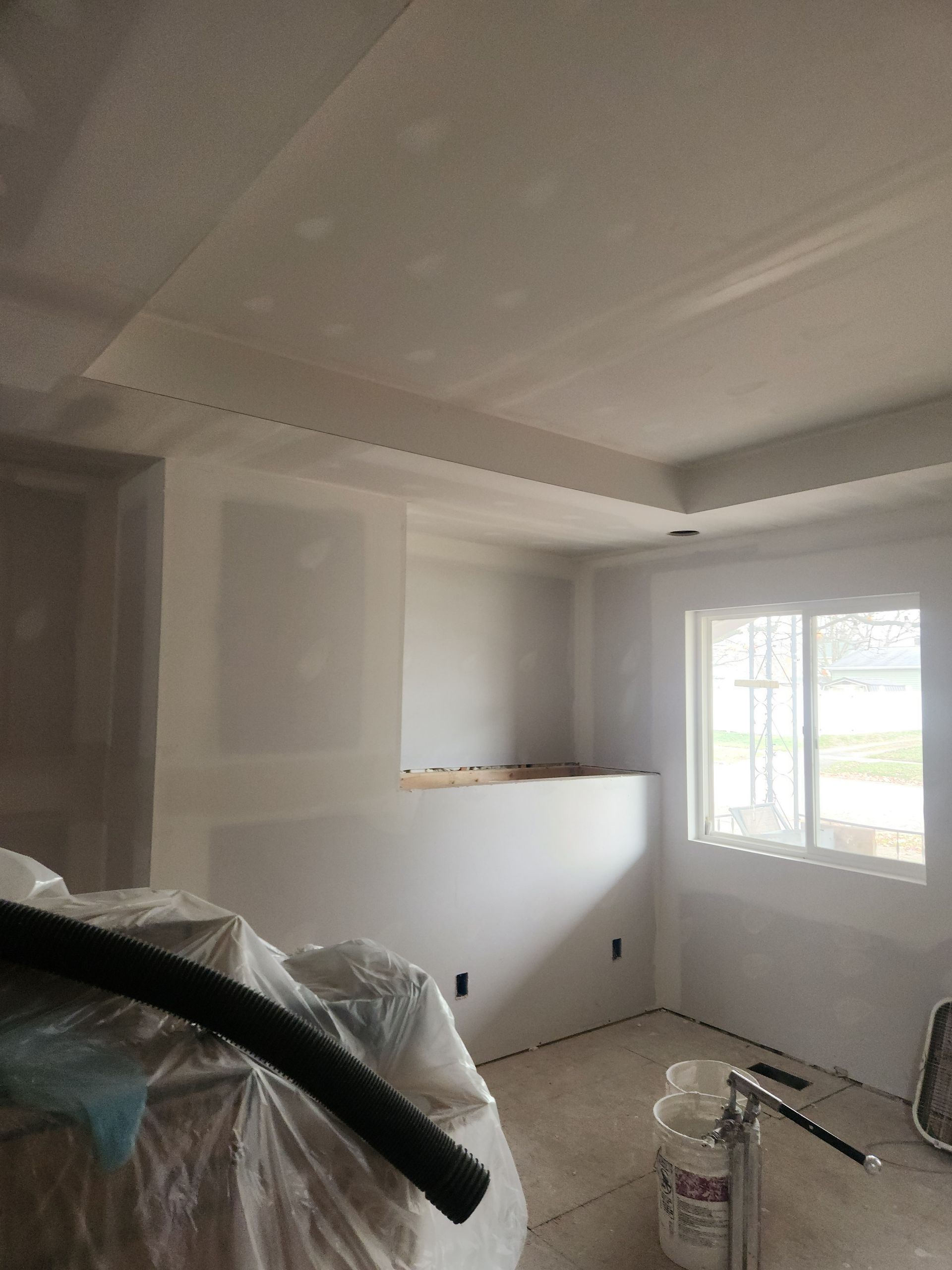 Interior room under construction; drywall walls, ceiling with recessed section, window, covered objects.