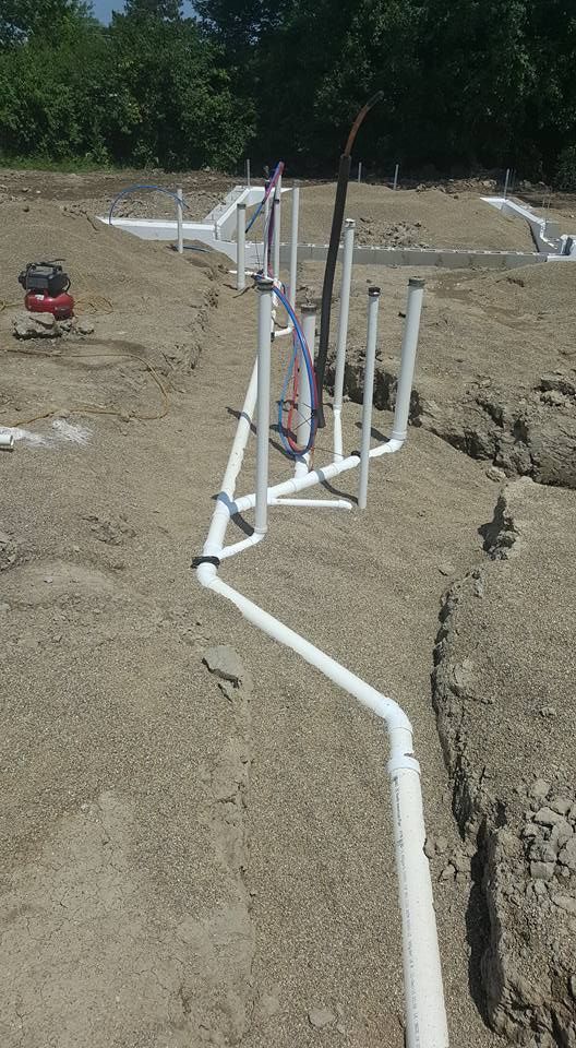 Plumbing pipes in trenches on a construction site, with dirt, gravel, and trees in the background.