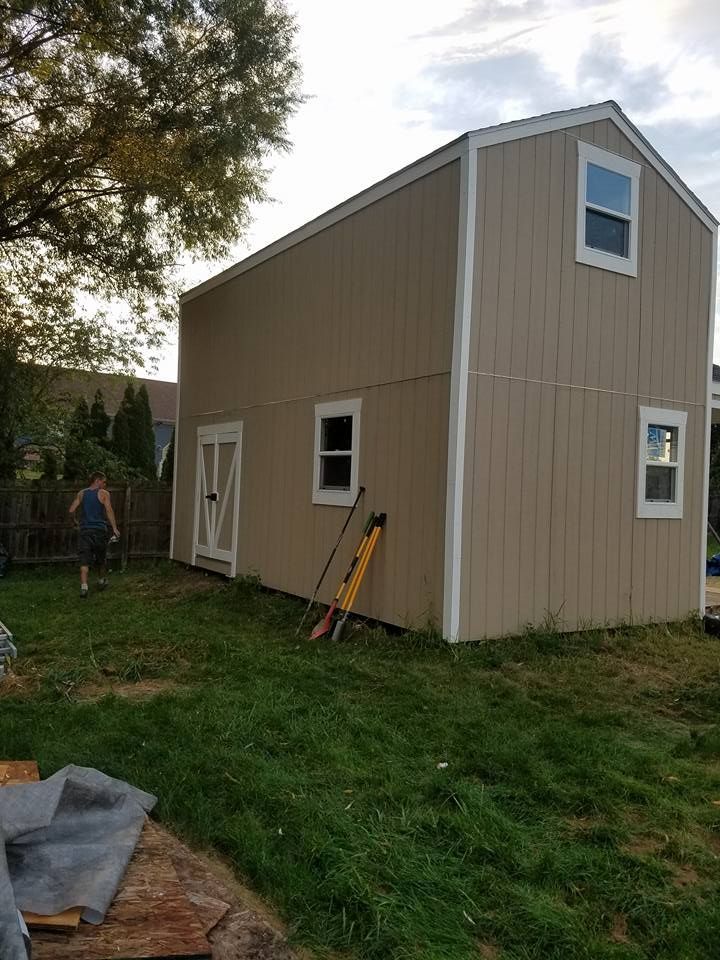 Tan two-story shed in a yard; a person stands near the shed, tools on the ground, cloudy sky.