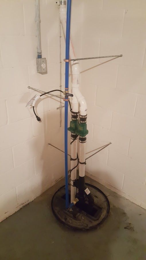 Sump pump system in a basement corner; black pump, white pipes, blue pipe, gray basin.