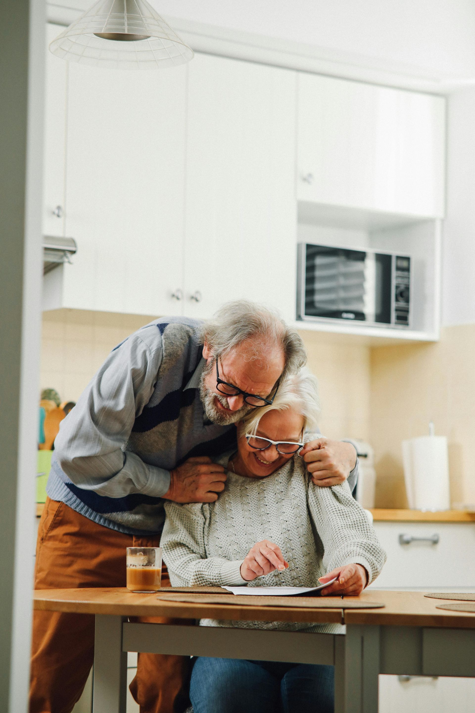 An older couple smiles while looking at a document together at a wooden dining table in a bright kitchen.