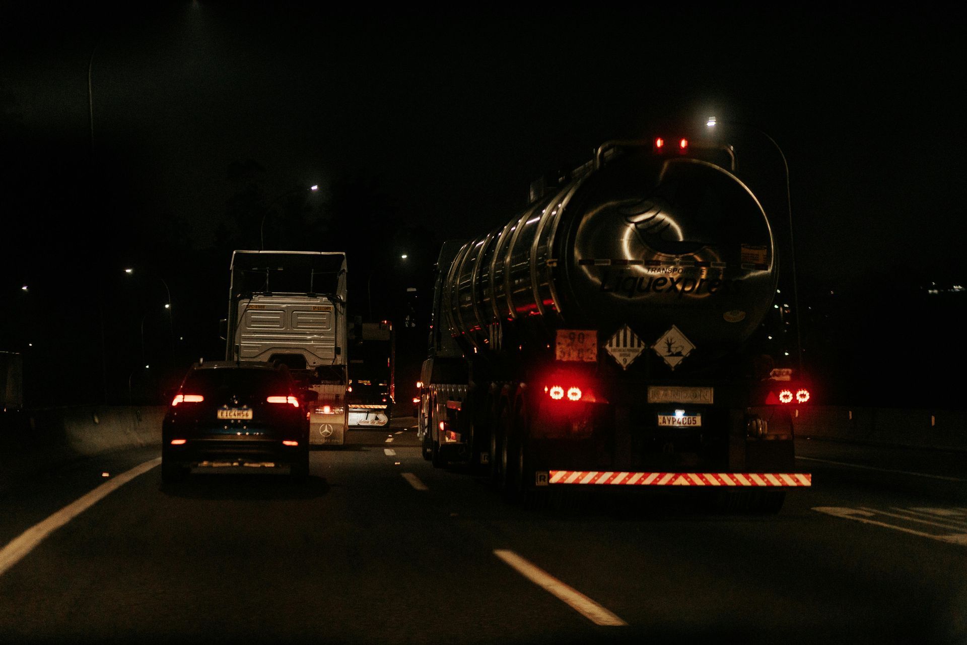 A nighttime road scene featuring a large tanker truck carrying hazardous materials alongside other vehicles.