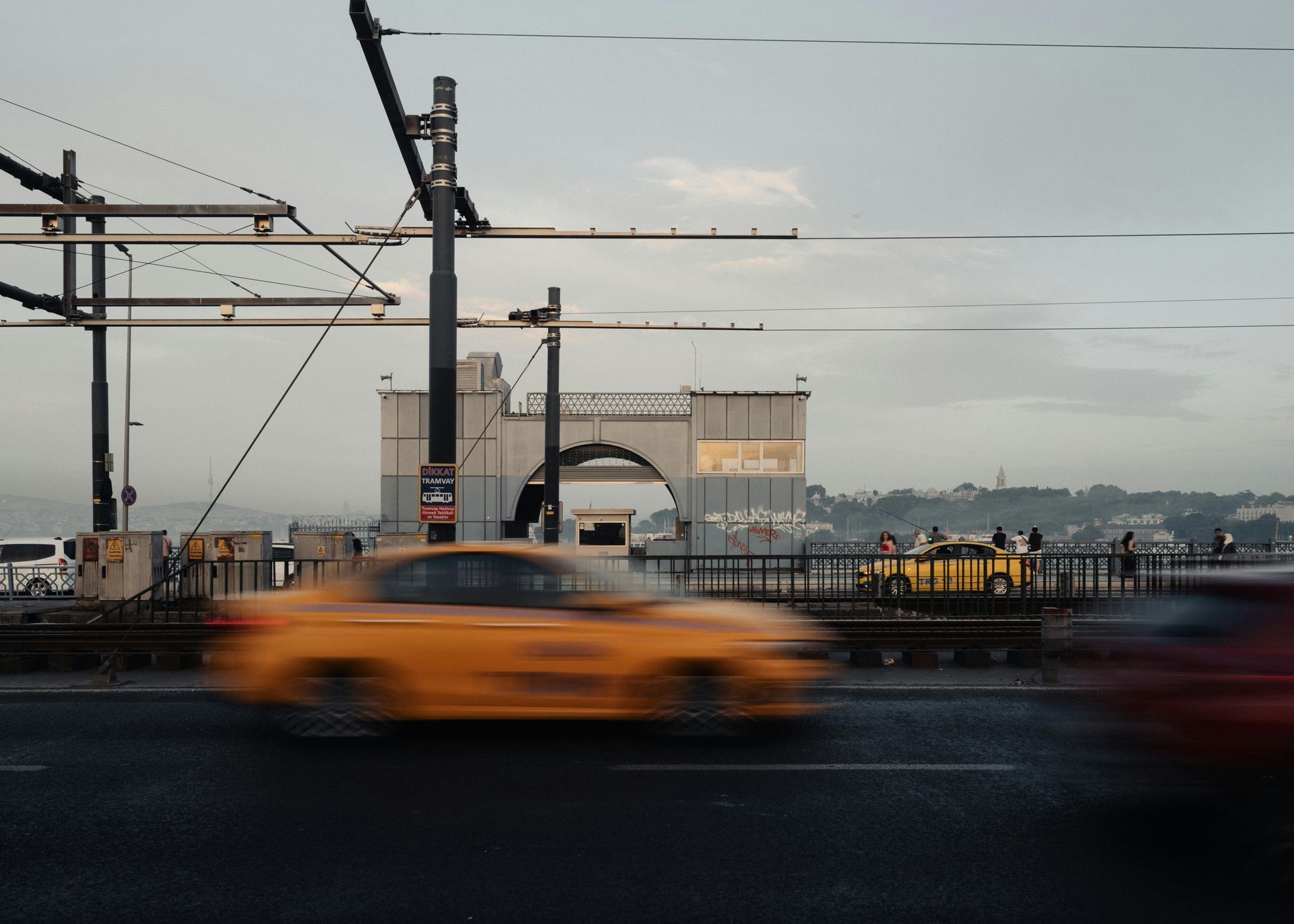A blurred yellow taxi speeds along a city bridge with a large stone arch structure in the background under a cloudy sky.