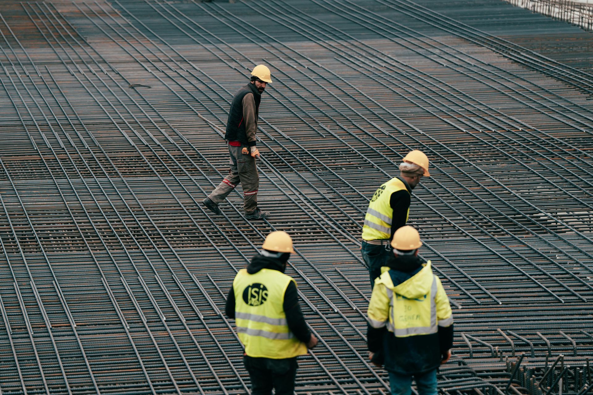 Four construction workers in safety gear walk across a large grid of steel rebar on a construction site.