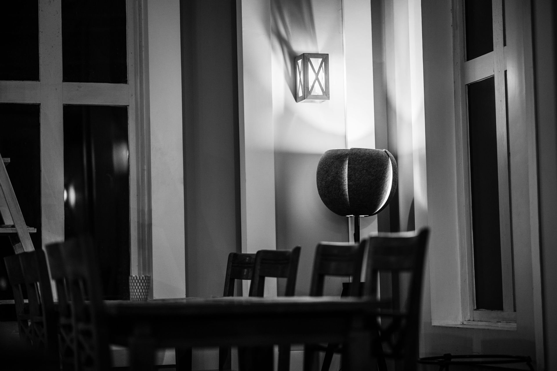 A black and white interior view of a dining room with a table, chairs, a wall sconce, and large window frames.