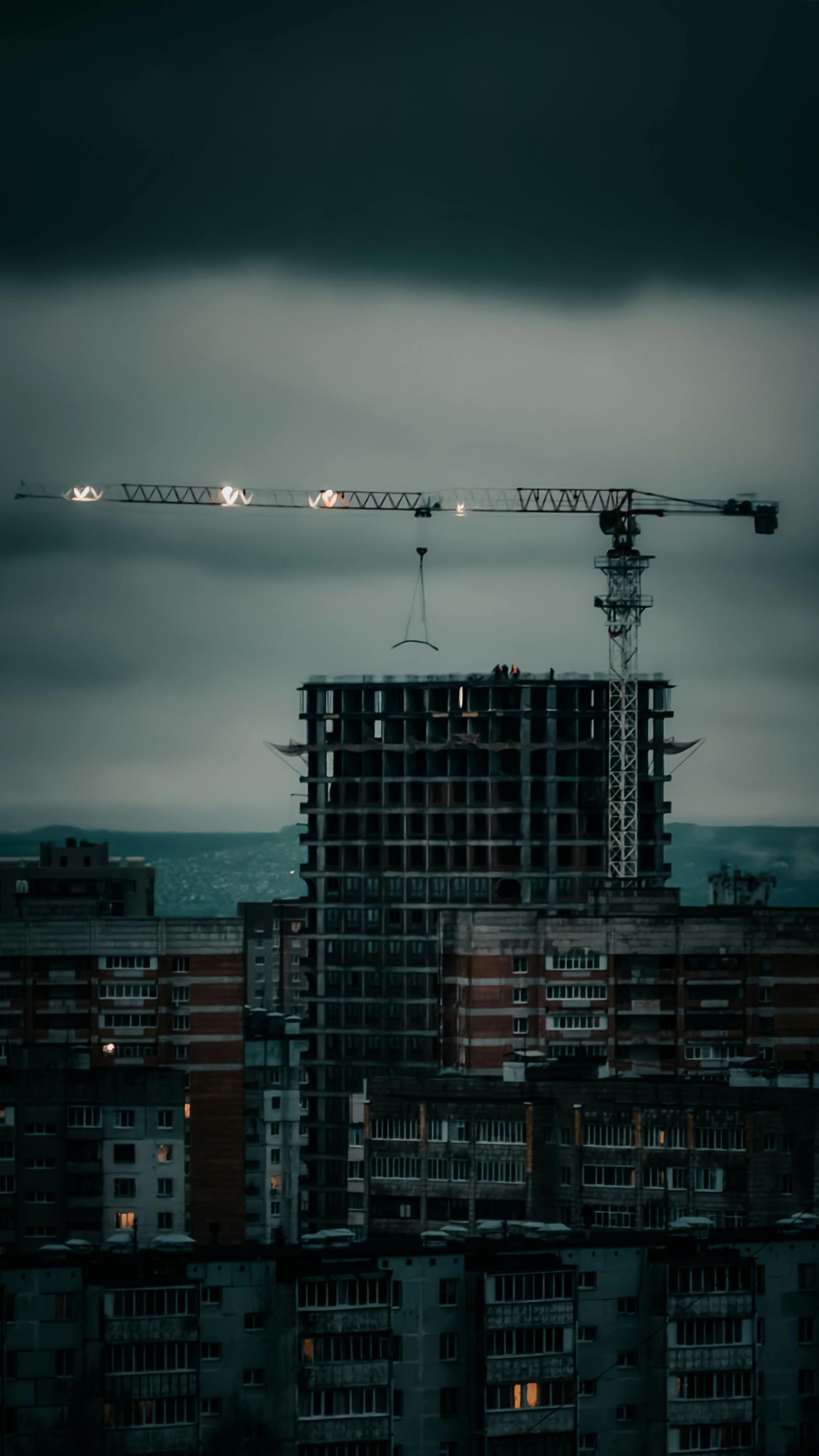 A tall construction crane looms over a dark, partially built concrete skyscraper under a moody, overcast sky.