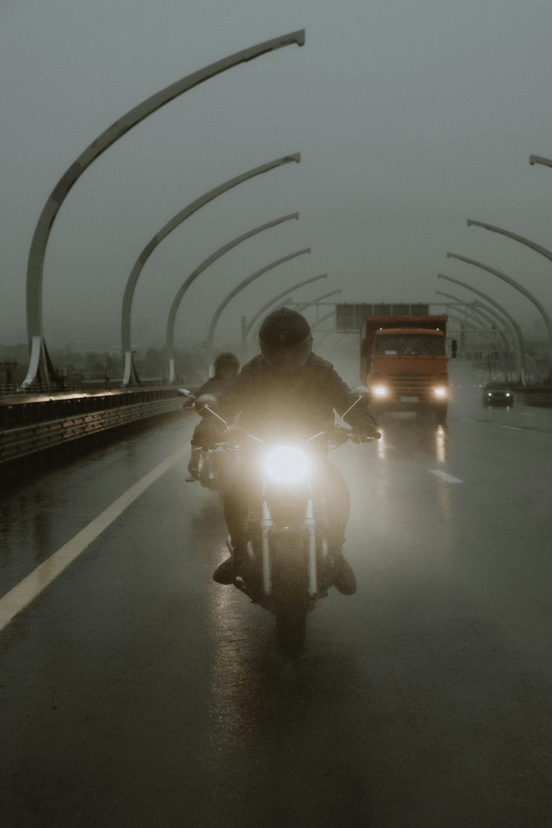 A motorcyclist rides toward the camera on a wet highway lined with curved streetlights under a cloudy, overcast sky.