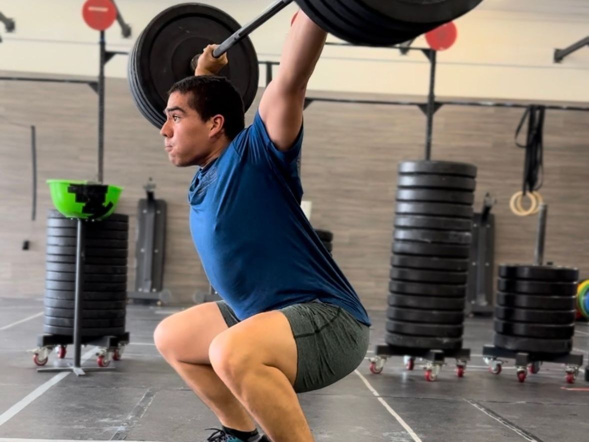 A man is squatting with a barbell over his head in a gym.
