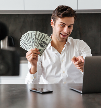 Man filming a vlog. He sits at a desk with a laptop, surrounded by cameras and a ring light. He smiles.