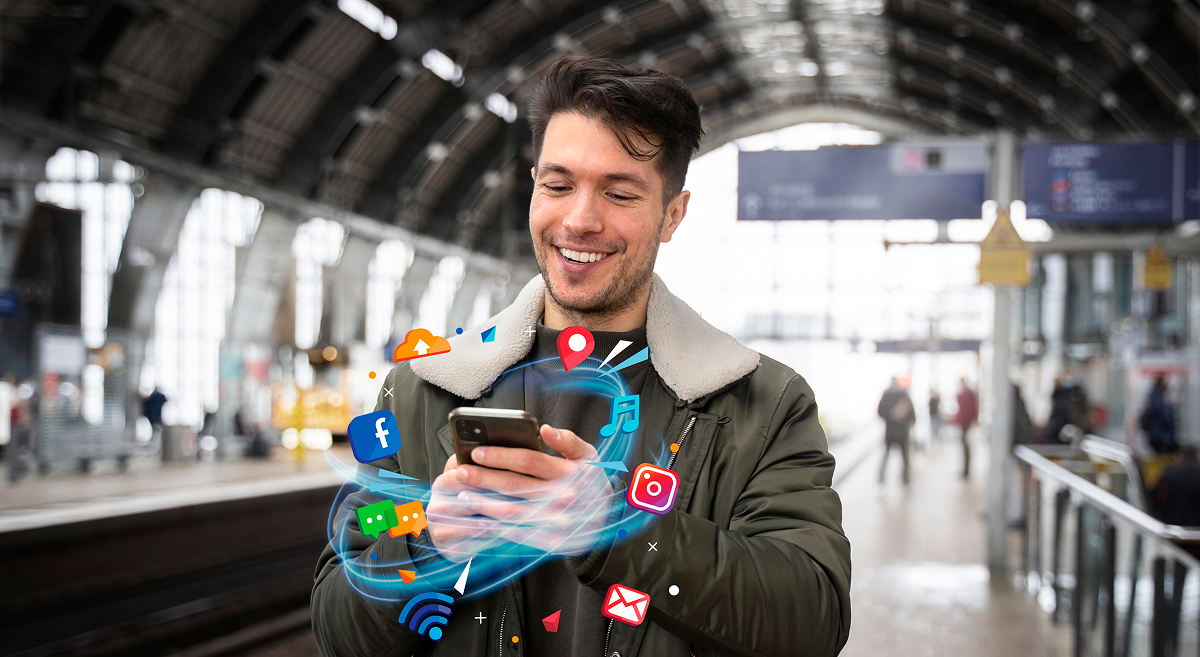 Man smiles at smartphone, surrounded by social media icons, at a train station.