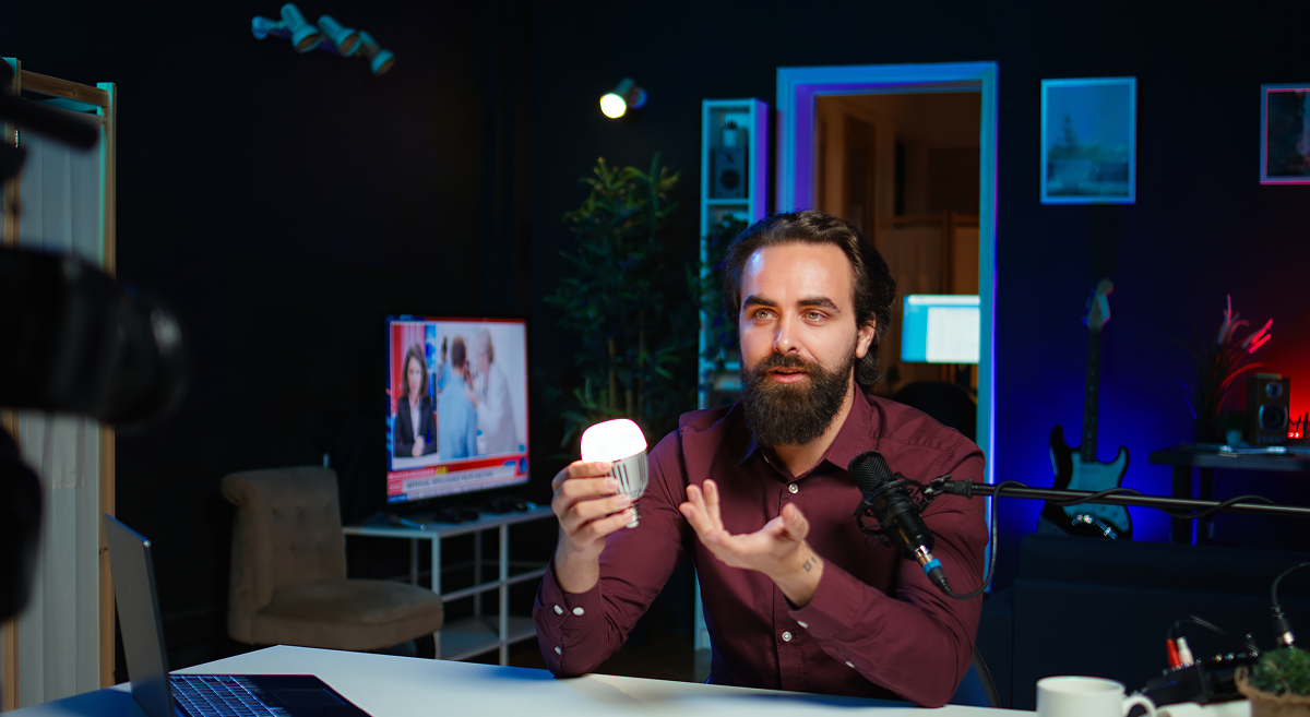 Man with beard holding a small light in a dark room with studio lights and a TV in the background.
