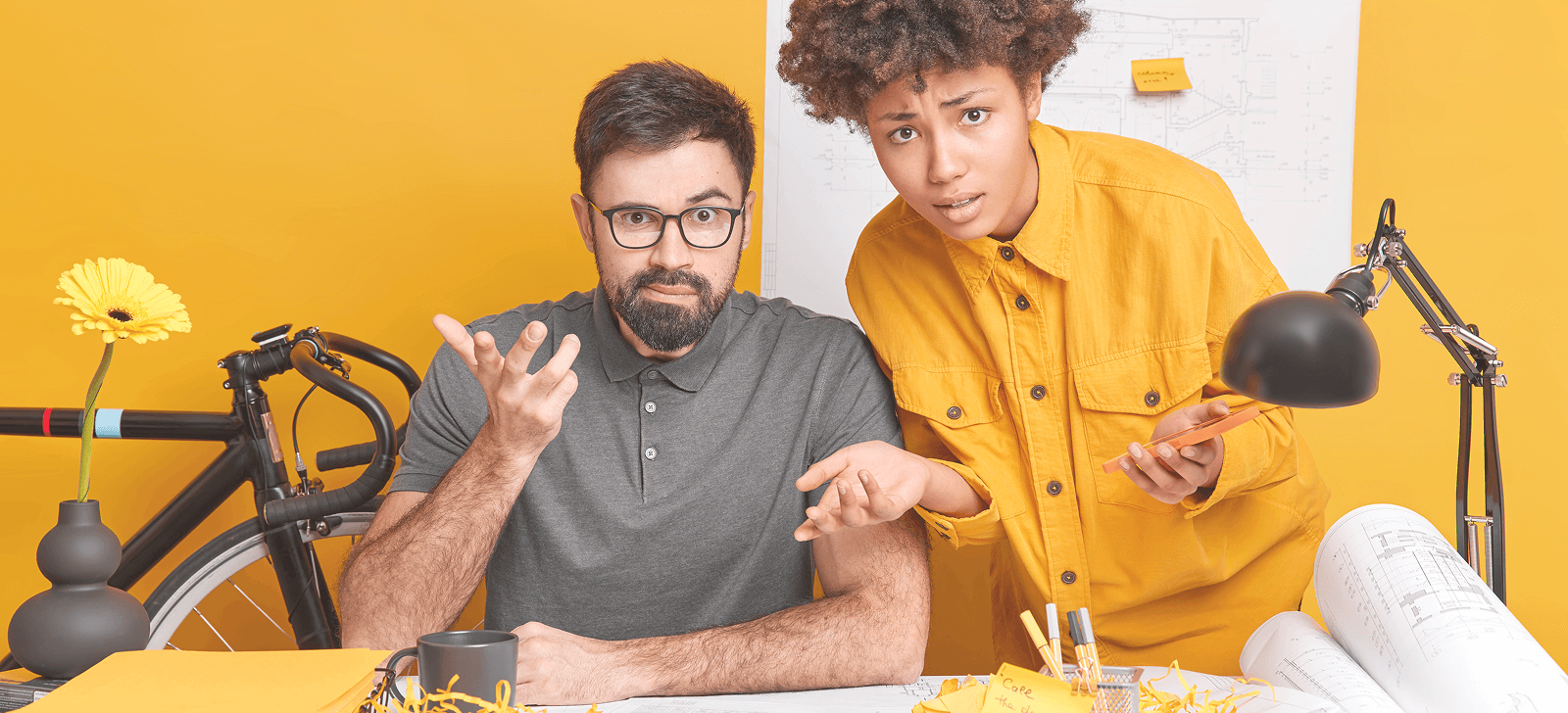 Two people looking puzzled at a desk. The background is yellow, there is a bicycle.