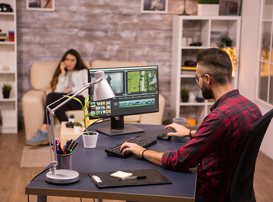 Man color-correcting video footage on dual monitors at a desk; he's in a dark room with a coffee cup.