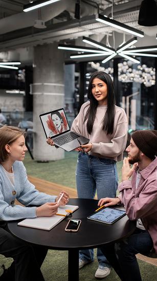 A woman presents a laptop with a design to two seated colleagues in a modern office setting.