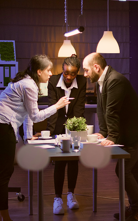 Three people in suits at a desk, in discussion. Woman gesturing, others looking on; office setting.