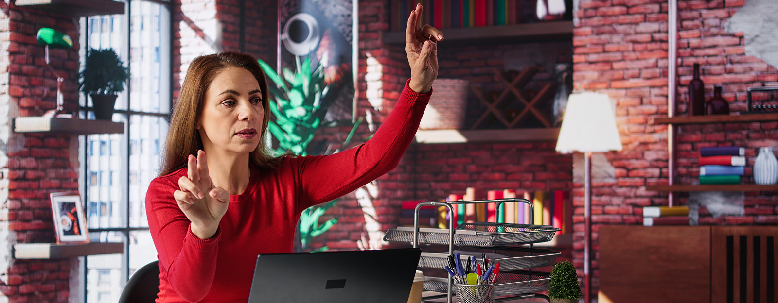 Woman in red top gesturing with hands in an office with brick wall and laptop.