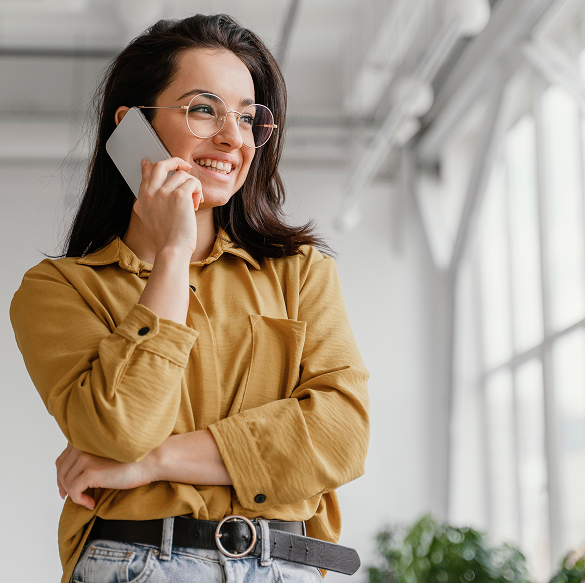 Woman with glasses smiles while on phone, standing near a window. She wears a yellow shirt and black belt.