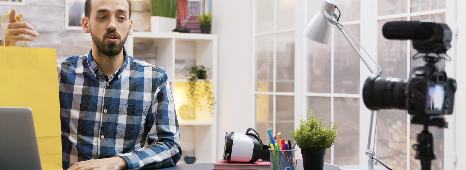 Man with beard in blue plaid shirt filming a video in a home studio. Camera and lamp in foreground.