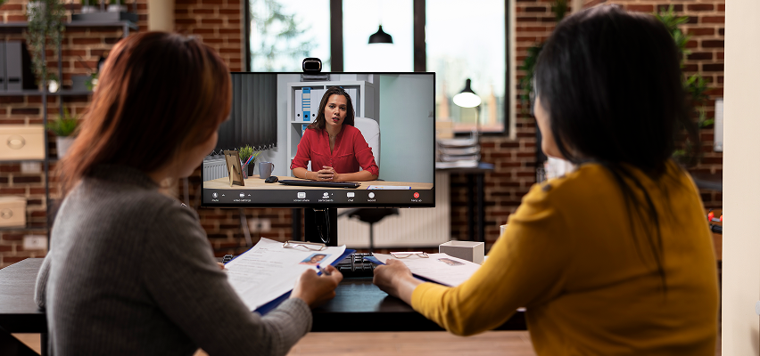 Two women on a video call in an office. The woman on the screen wears a red shirt.