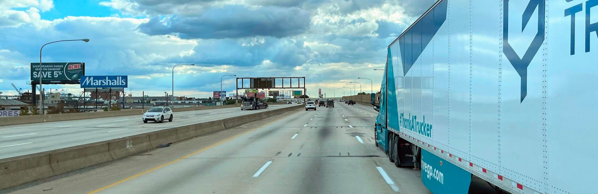 A semi-truck on a highway, passing other vehicles under a cloudy sky. Billboard signs visible.