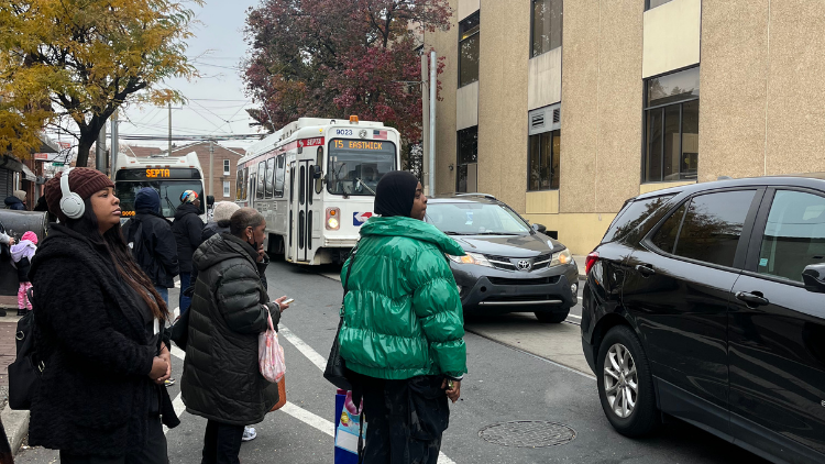 People waiting at a trolley stop; a trolley and a car are present. Overcast day.