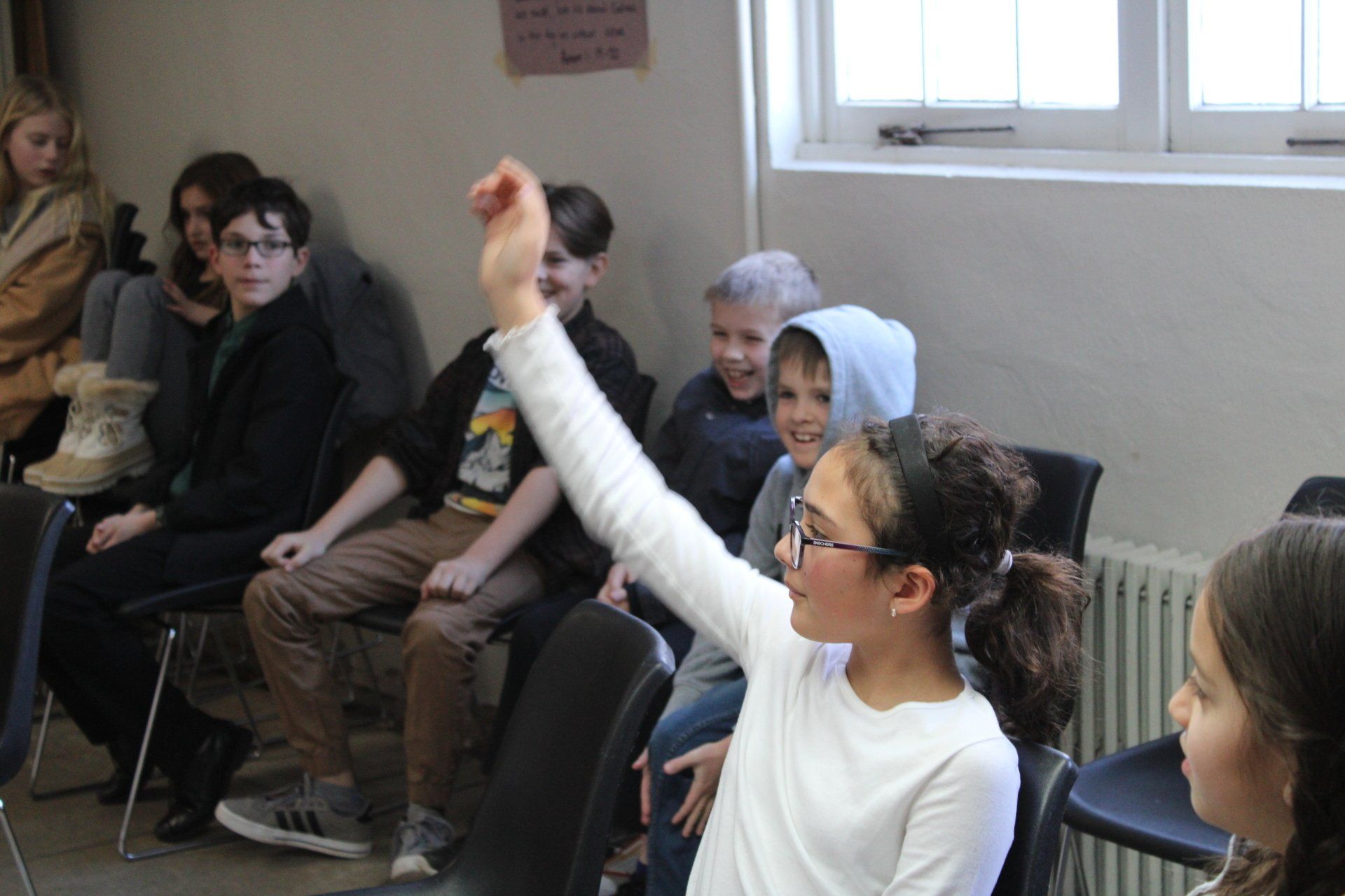 A group of children are sitting in chairs and a girl is raising her hand to answer a question.