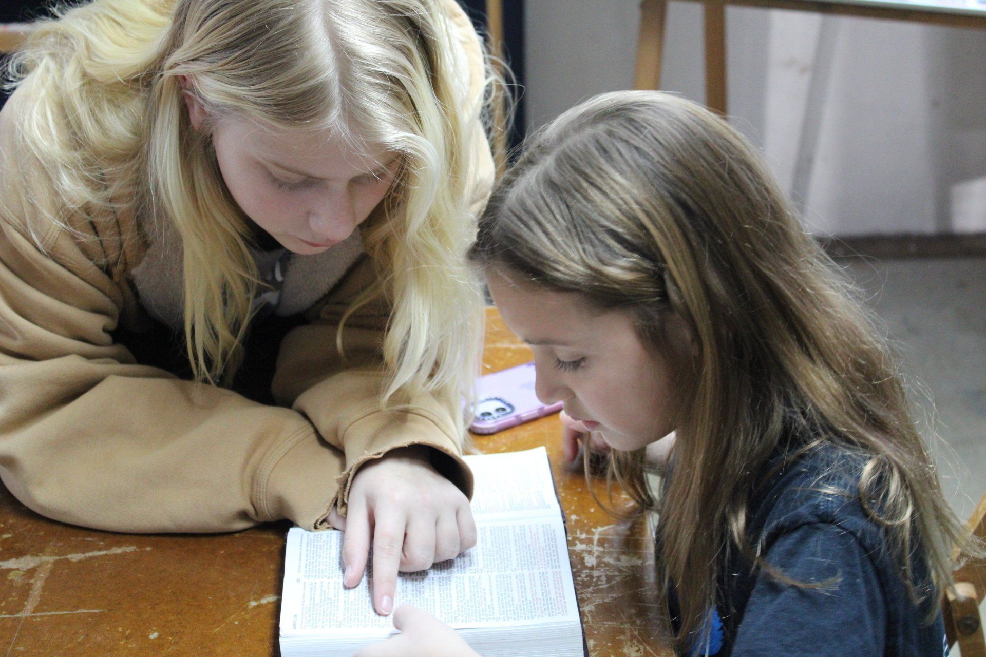 Two young girls are sitting at a table reading a book.