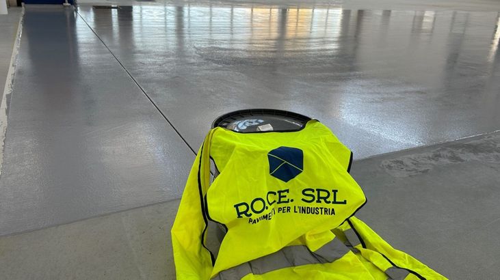 Giubbotto di sicurezza giallo con logo ROCE SRL, accanto al pavimento grigio lucido appena verniciato in un edificio.