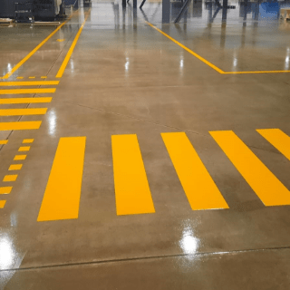 Attraversamento pedonale e linee dipinte di giallo su un pavimento in cemento grigio lucidato in un ambiente industriale.