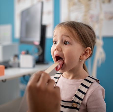 Child opening mouth for a tongue exam in a bright clinic, with a doctor’s hand holding a tongue depressor.
