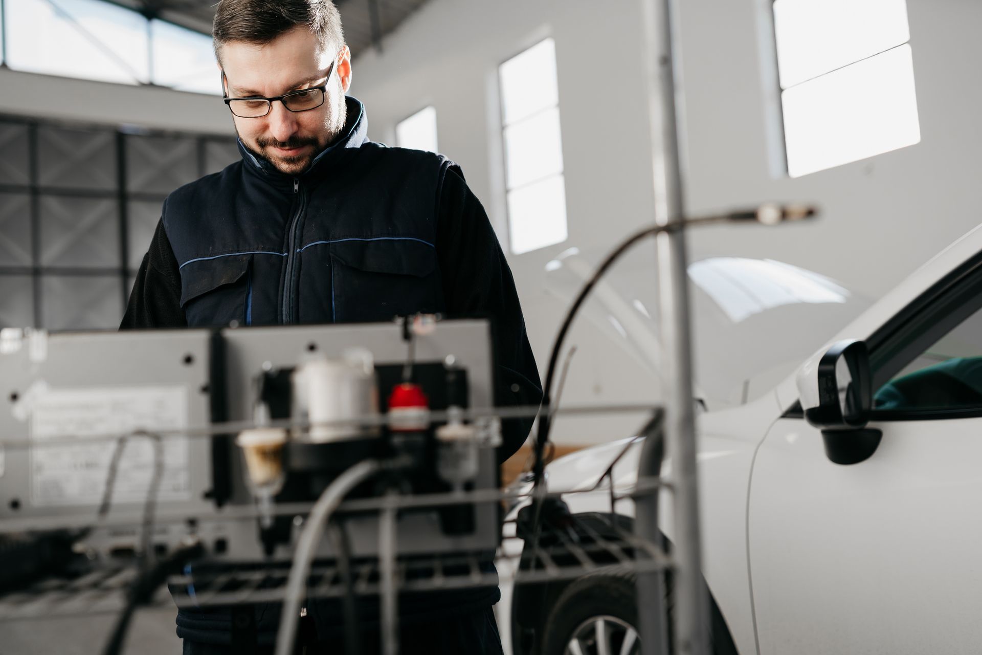 Mechanic in glasses working on a car in a garage, looking at a diagnostic machine.