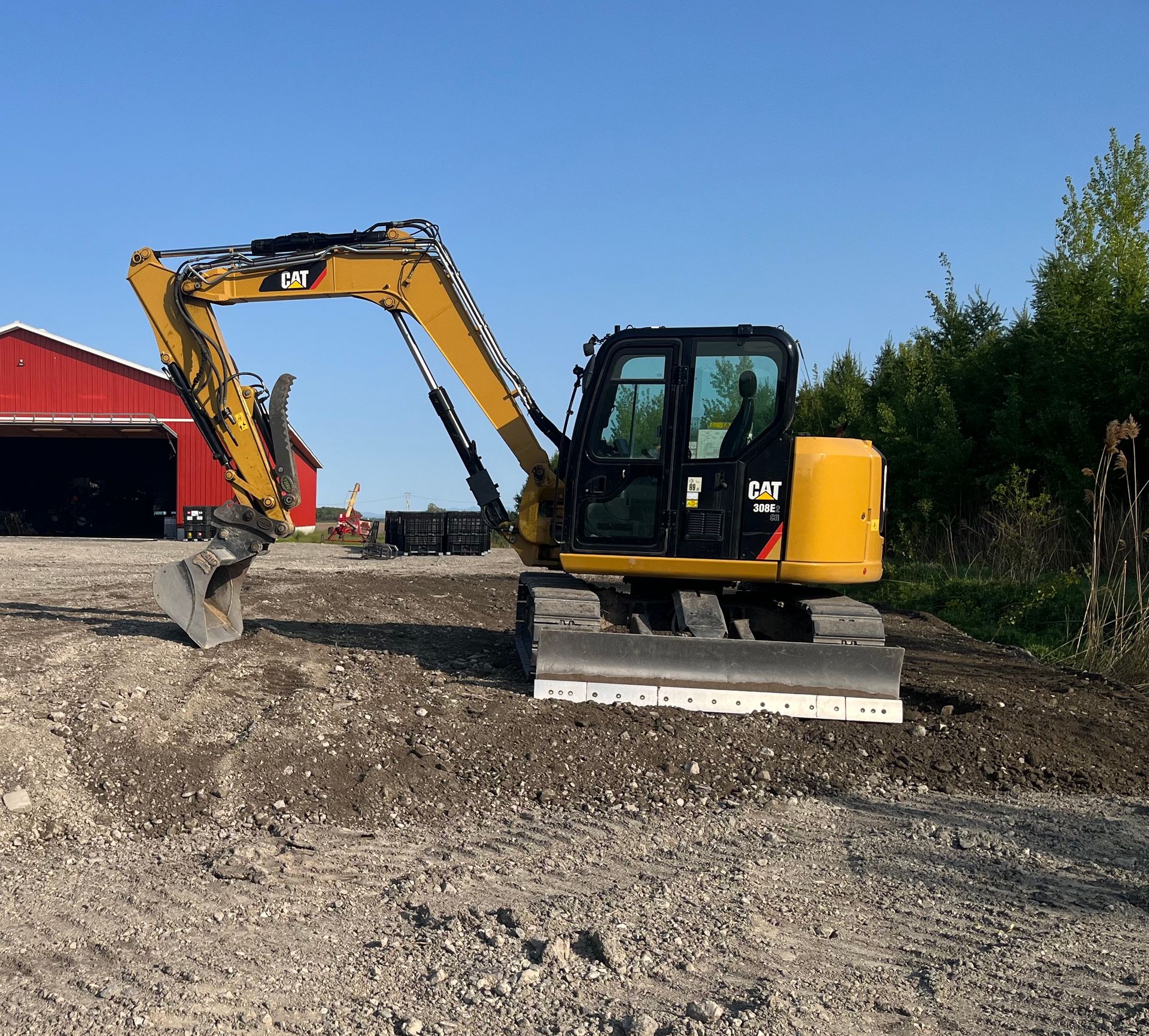 Une pelleteuse jaune sur du gravier devant une grange rouge et des arbres sous un ciel bleu.