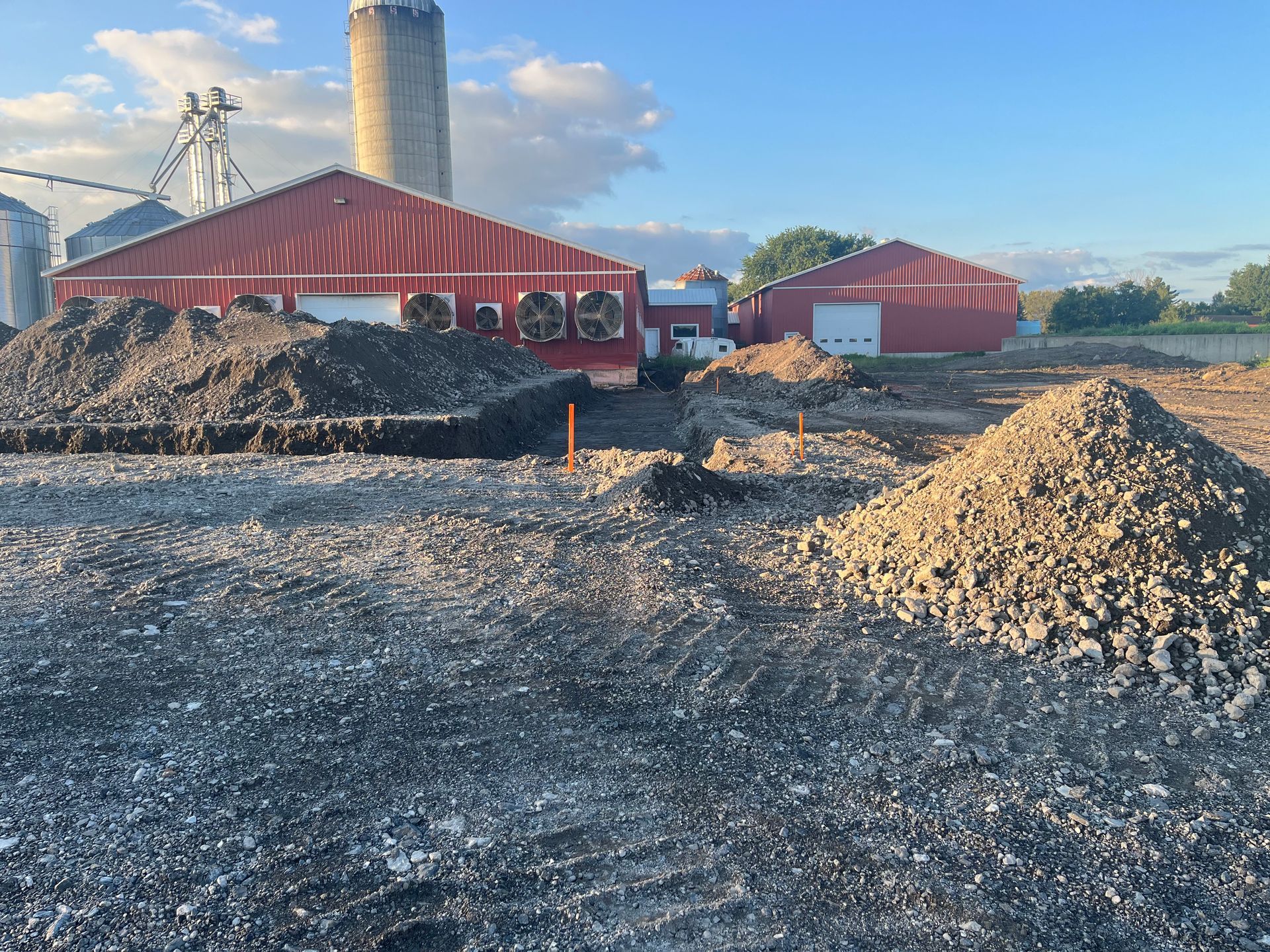 Bâtiments agricoles aux toits rouges en construction ; tas de terre au premier plan ; silo à l'arrière-plan.